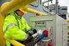 The image shows a man dressed in a yellow high vis connecting a vessel to shore power