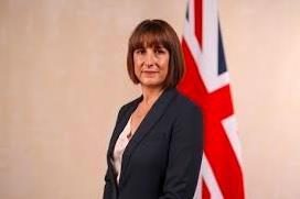 The image shows Rachel Reeves dressed in a grey suit standing in front of the Union Jack flag