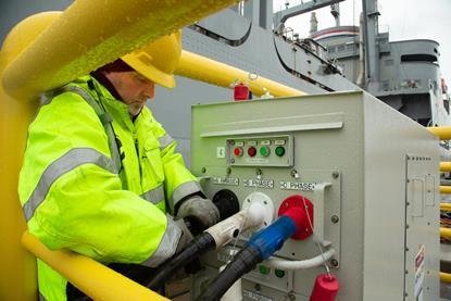 The image shows a man dressed in a yellow high vis connecting a vessel to shore power