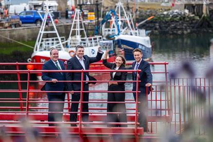 Four members of Eyemouth Harbour Trust standing on a red gangway holding aloft the harbour's masterplan