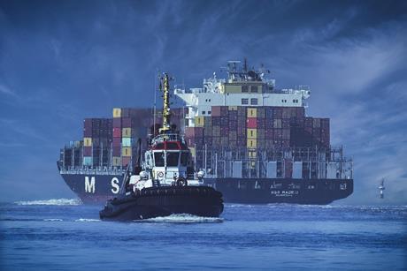 The image shows a container ship in the background with a tug in the foreground